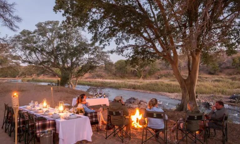 Dîner en plein air près d'une rivière lors d'un safari à pied Kruger, avec des personnes assises autour d'un feu de camp sous des arbres.